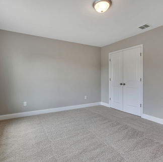 Empty bedroom with light gray walls, beige carpeting, and a large window allowing natural light. A double-door closet with white paneled doors is centered on one wall, and a flush-mount ceiling light fixture is installed above.