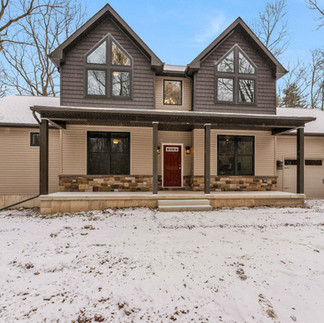 Front exterior view of a two-story house in East Stroudsburg, PA with dark shingle gables, tan siding, a covered porch with stone accents, red front door, and attached two-car garage, set in a snowy yard with leafless trees.
