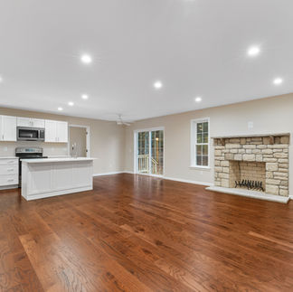 Interior view of an unfurnished open-concept kitchen and living room. The space has brown hardwood floors, beige walls, and recessed lighting. The kitchen has a center island with a gooseneck faucet, stainless range and microwave, and a refrigerator. There are sliding glass doors with a ceiling fan nearby to the right of the kitchen. A stone fireplace with a wood mantel sits between two tall windows.
