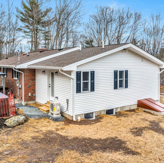 Exterior side angled view of a single-story house in Scotrun, PA with beige siding one half of the house and brick exterior on the other half, and black shutters. A set of wooden stairs leads to a back door, with a small outdoor unit and utility features beside it. There is a gazebo structure in the backyard.