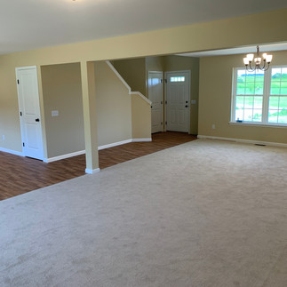 Interior view of an unfurnished open-concept living area and entry way. The living room has light gray carpeting leading into a dining nook with a multi-light chandelier beneath a beam and column. The room has beige walls with white trim, a large front window, wood-look flooring at the entry and hallway, white paneled doors, and a front door beside a stairway that leads upstairs.