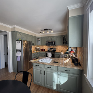 Interior view of a kitchen with sage green cabinets, stainless steel refrigerator and microwave, and a granite peninsula countertop with mail and small appliances; tan tile backsplash and large windows on the right provide natural light.