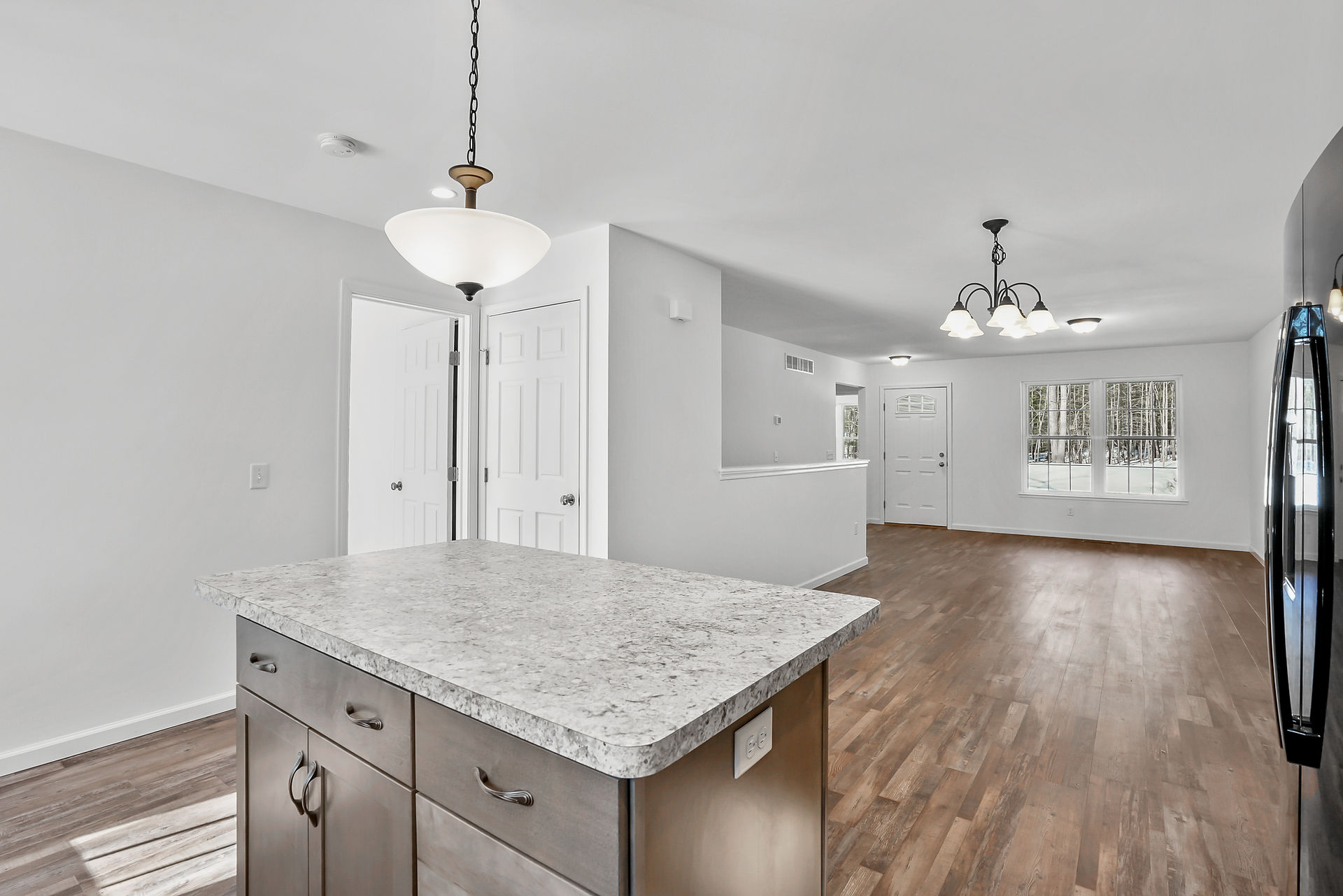 Open-concept kitchen and unfurnished living space with wood-style flooring and white walls. View from behind a granite-look island with storage cabinets shows a living area with a central front door, large window, and multiple light fixtures including a chandelier and ceiling lights. Two interior doors are visible near the kitchen.