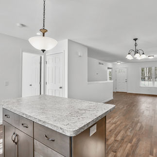 Open-concept kitchen and unfurnished living space with wood-style flooring and white walls. View from behind a granite-look island with storage cabinets shows a living area with a central front door, large window, and multiple light fixtures including a chandelier and ceiling lights. Two interior doors are visible near the kitchen.