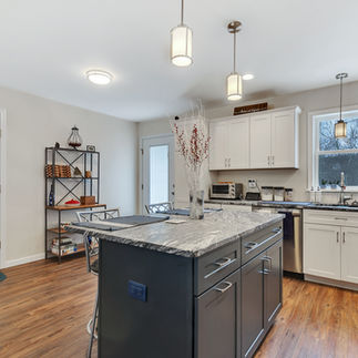 Interior view of finished kitchen with a large island with a dark base and granite countertop, white cabinets and stainless steel appliances. Pendant lights hang above the island and sink, and a shelving unit stands against the back wall. A window above the sink allows natural light.