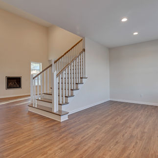 Open-concept unfurnished living area with light wood flooring, white walls, recessed lighting, and a staircase leading upstairs with wooden steps and white railing. The room has windows providing natural light and a built-in fireplace.