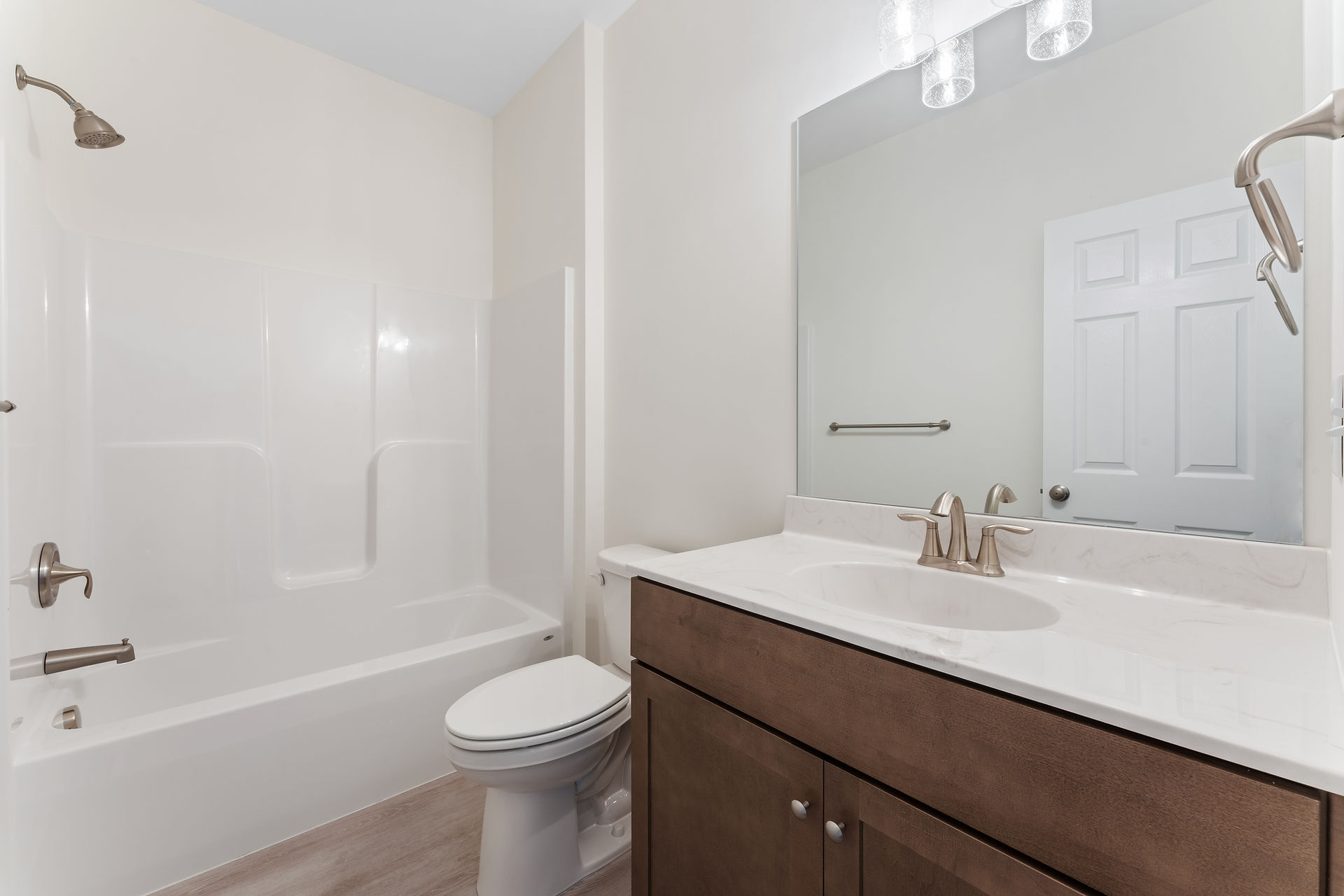 Finished bathroom with a single-sink vanity with dark wood cabinetry a white quartz countertop, white bathtub and shower combination, a large frameless mirror above the sink, and contemporary light fixtures above. Walls are painted a neutral tone with light wood-look flooring.