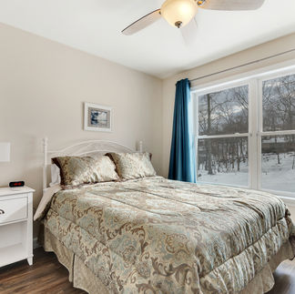 Furnished bedroom with a queen-sized bed with a patterned comforter and matching pillows, next to a large window with blue curtains. The room has a white nightstand with a lamp and digital clock, wood-style flooring, and a ceiling fan with a light fixture.