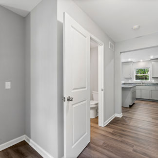Interior hallway leading to a kitchen with gray cabinets, granite countertops, and stainless steel appliances. The space has light gray walls, wood-style vinyl plank flooring, and recessed lighting. A partially open door reveals a small bathroom with a visible toilet, and a carpeted staircase is to the left. Natural light enters through a window above the kitchen sink.