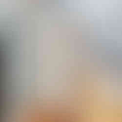 Interior view of a bathroom with light gray walls, wood plank floors, and a vaulted ceiling. There is a partially open white door with light wood trim in the middle of the room that leads into a hallway. On the left is a walk in shower with white subway tile walls and a built in niche, brown hexagonal tile floors, and glass paneled sliding doors with black frames. On the right is a wood slate vanity with double white vessels sinks with black water spouts. A black towel ring with a black and white striped towel on on the back wall and a white outlet is placed in between the two sinks.