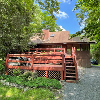 Exterior front view of a two-story brown wooden house in Pocono Lake, PA with a large red wooden deck and stairs, surrounded by trees and greenery, with a gravel driveway on the right side.