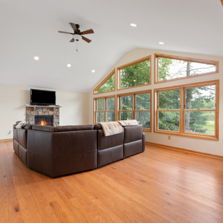 Furnished living room with hardwood floors, vaulted ceilings with recessed lighting and a ceiling fan, and a wall of large windows offering views of trees and greenery. A stone fireplace with an active fire and a wall-mounted television are on the back wall, with a large brown leather sectional sofa in the center of the room.