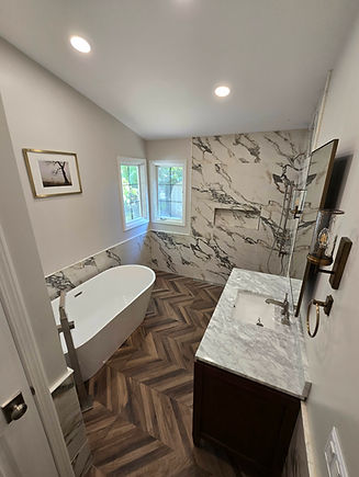 This close-up view of the completed bathroom remodel in West Bloomfield, Michigan highlights the sophisticated material palette and custom detailing. The walk-in shower features striking marble-look tile with a recessed niche, a frameless glass panel, and a modern rain and handheld shower system. The dark wood vanity with marble countertop adds warmth and contrast, while herringbone tile flooring ties the entire space together with elegance.