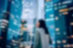 Uma mulher com um casaco cinzento está em pé numa paisagem urbana moderna ao entardecer, rodeada por arranha-céus altos de vidro com janelas iluminadas, olhando para cima com uma expressão contemplativa.