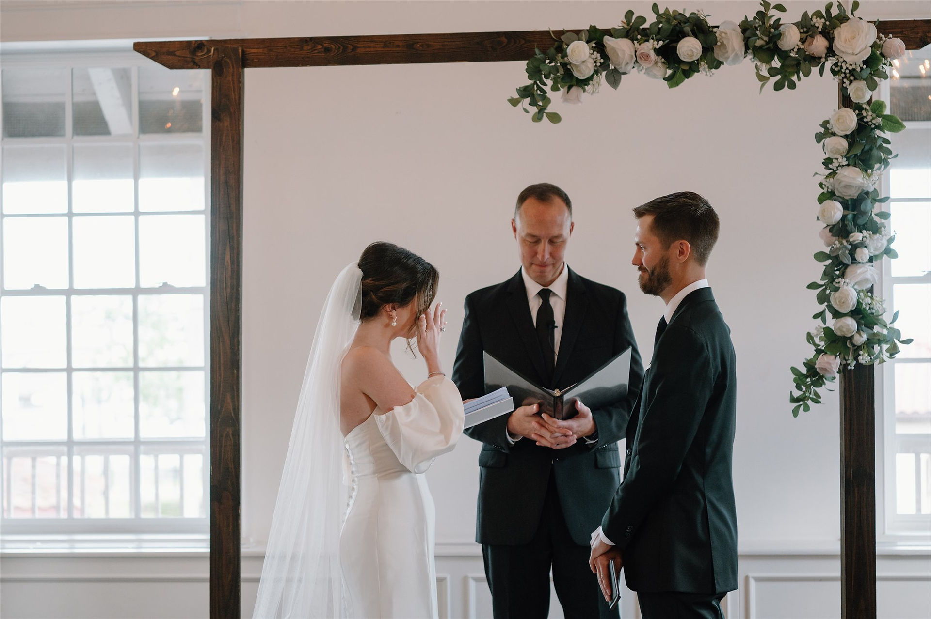 A bride and groom exchange rings during their wedding ceremony officiated by a man in a black suit at the white room