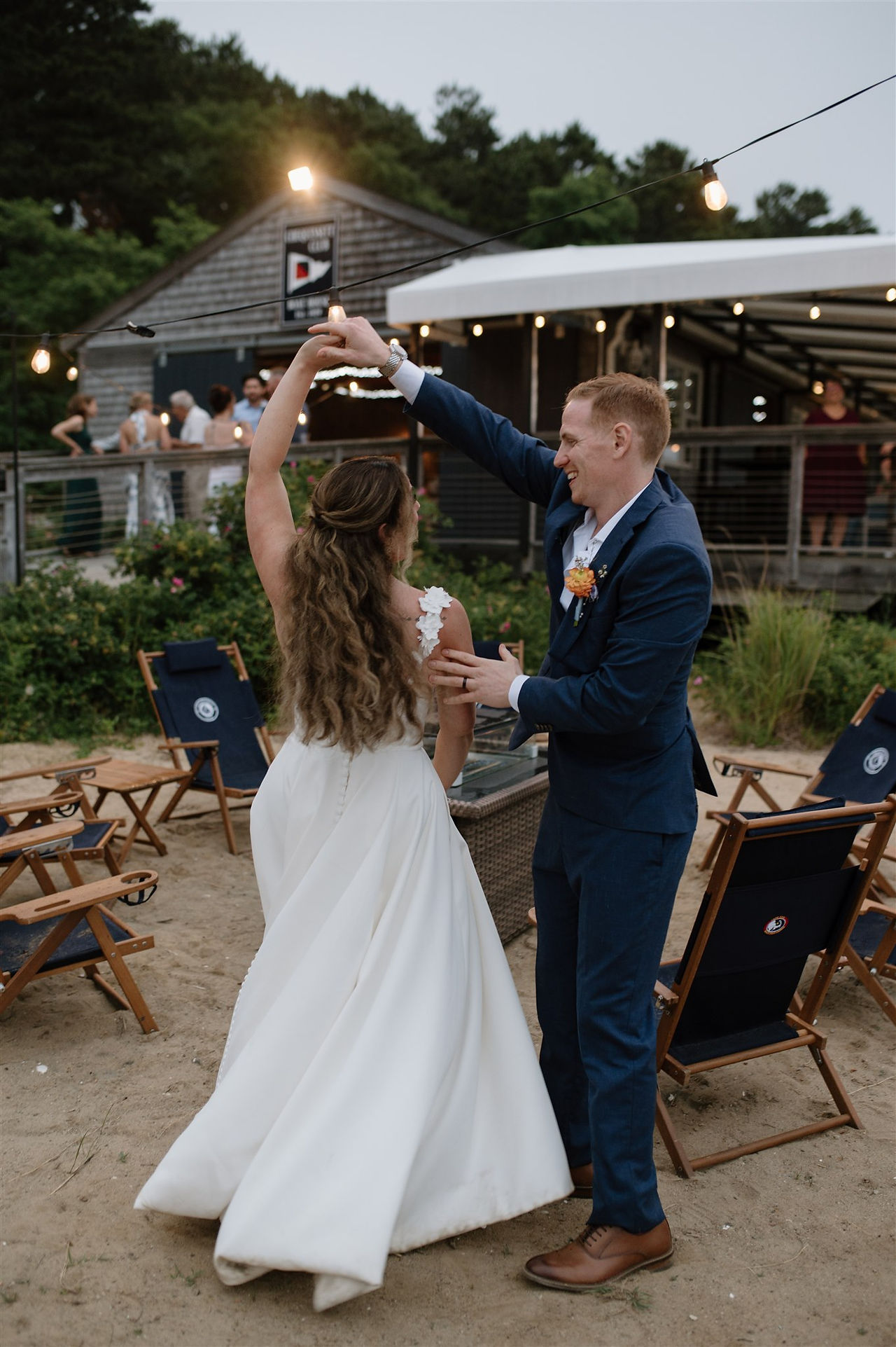 A bride and groom dance together outdoors on a sandy area, with a wooden building and string lights in the background.