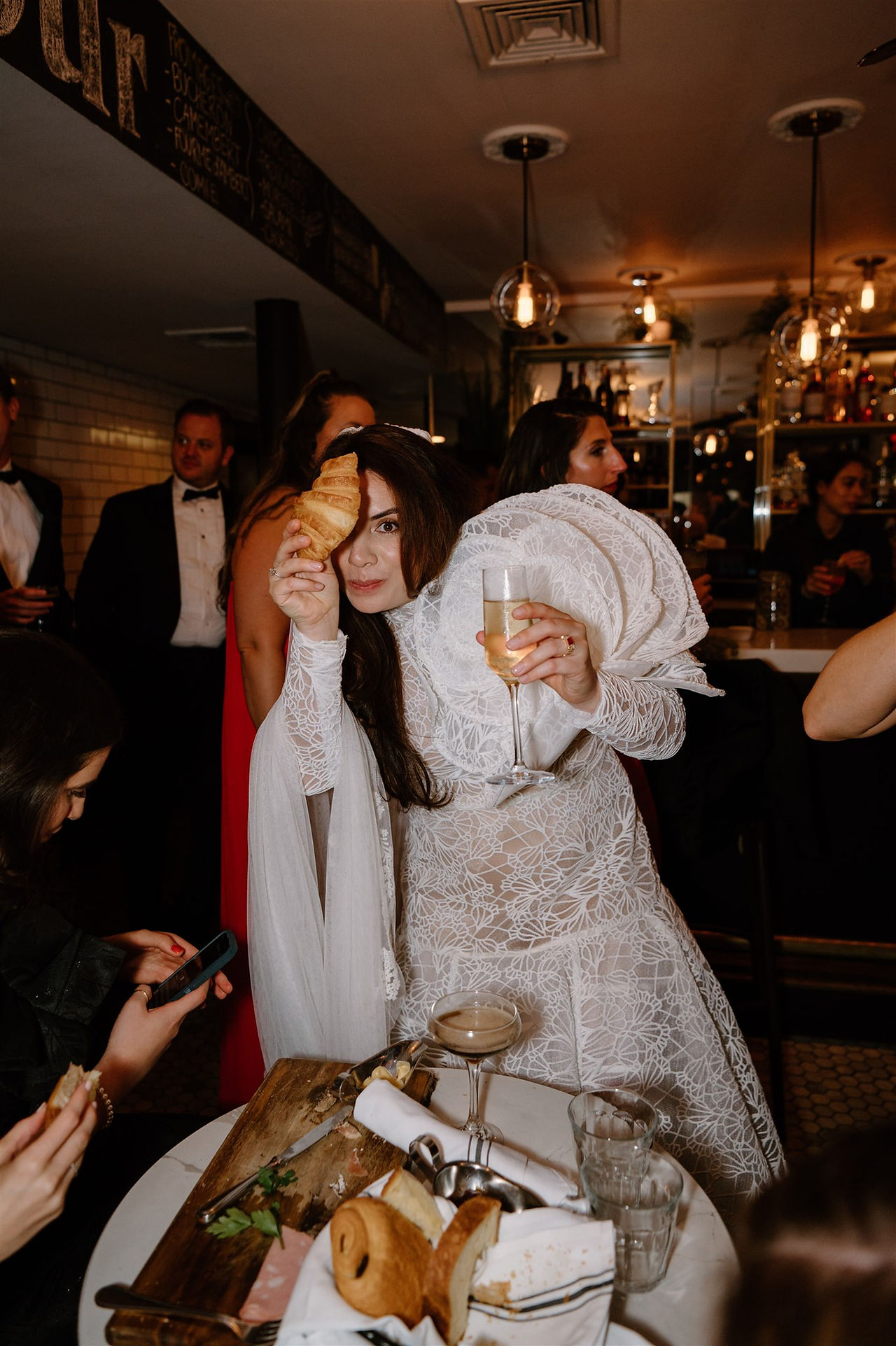 A person in a white lace outfit holds a croissant above their eye and a drink in their other hand at a gathering. Other attendees are interacting in the background.