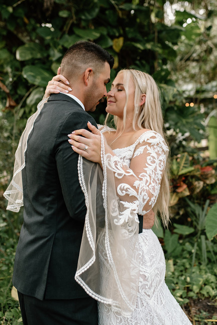 A bride and groom in formal attire embrace in a lush garden setting. The bride wears a long, white gown with lace details, and the groom is in a dark suit.