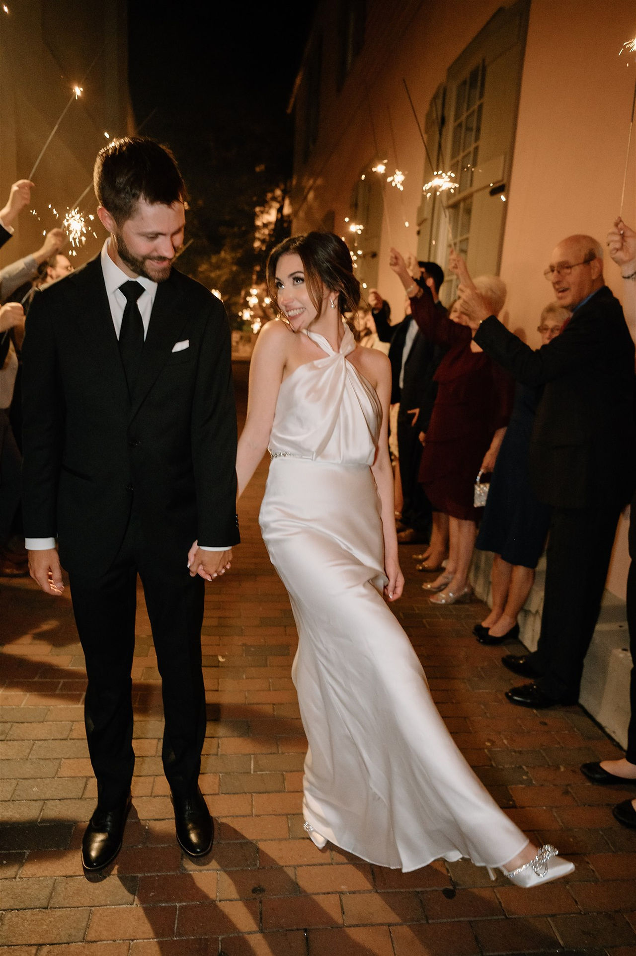 A couple, dressed formally in a black suit and a white dress, walk hand in hand through a crowd holding sparklers at night.