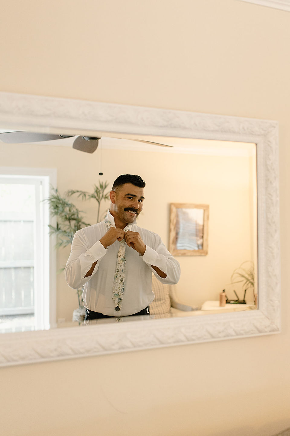 A man adjusts his tie while looking into a mirror in a warmly lit room.