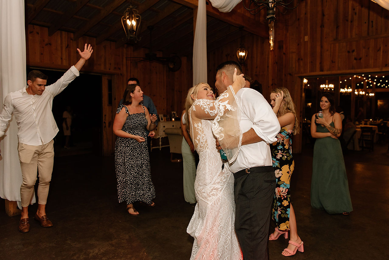 A bride and groom are dancing closely at a wedding reception. Guests in the background watch and smile.