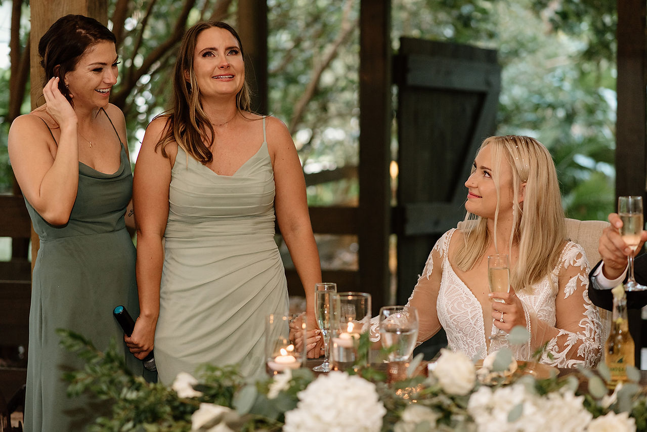 Three women celebrating indoors with drinks, one in a wedding dress and the others in green dresses, surrounded by flowers and candles.
