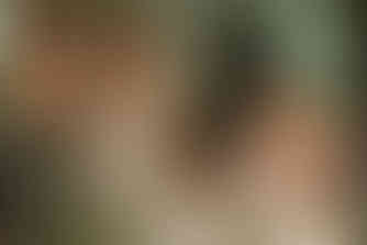 Three women celebrating indoors with drinks, one in a wedding dress and the others in green dresses, surrounded by flowers and candles.