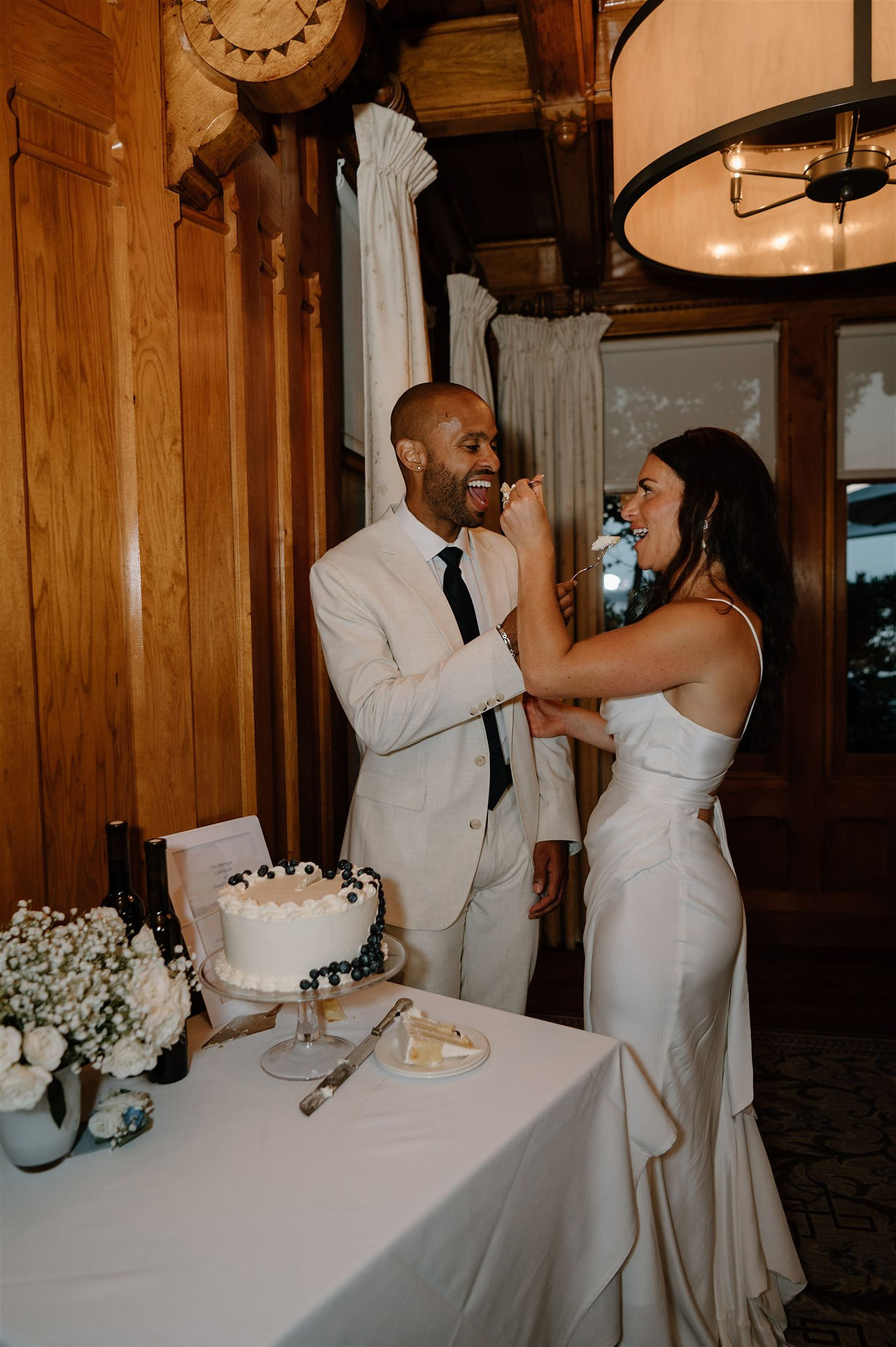 A bride and groom stand by a cake, with the bride feeding a piece to the groom indoors with wooden walls in the background.