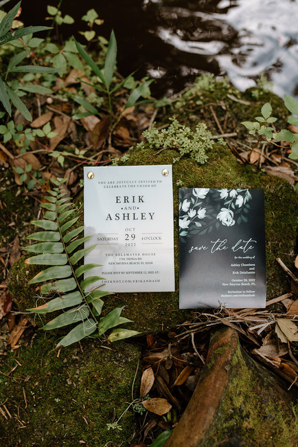Wedding invitation flat lay with shoes, white roses, eucalyptus, a perfume bottle, a wooden ring box, and hair accessories.