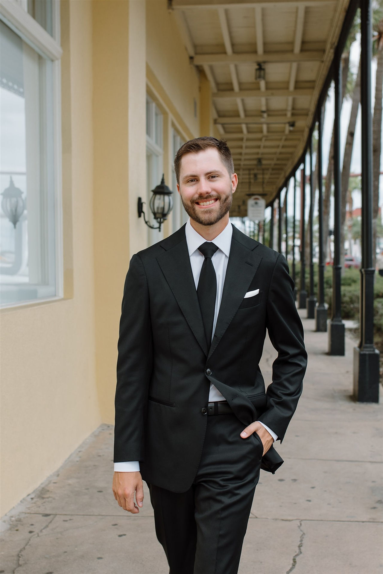 A smiling man in a black suit and tie walks down a corridor with columns and lanterns for his wedding at the white room