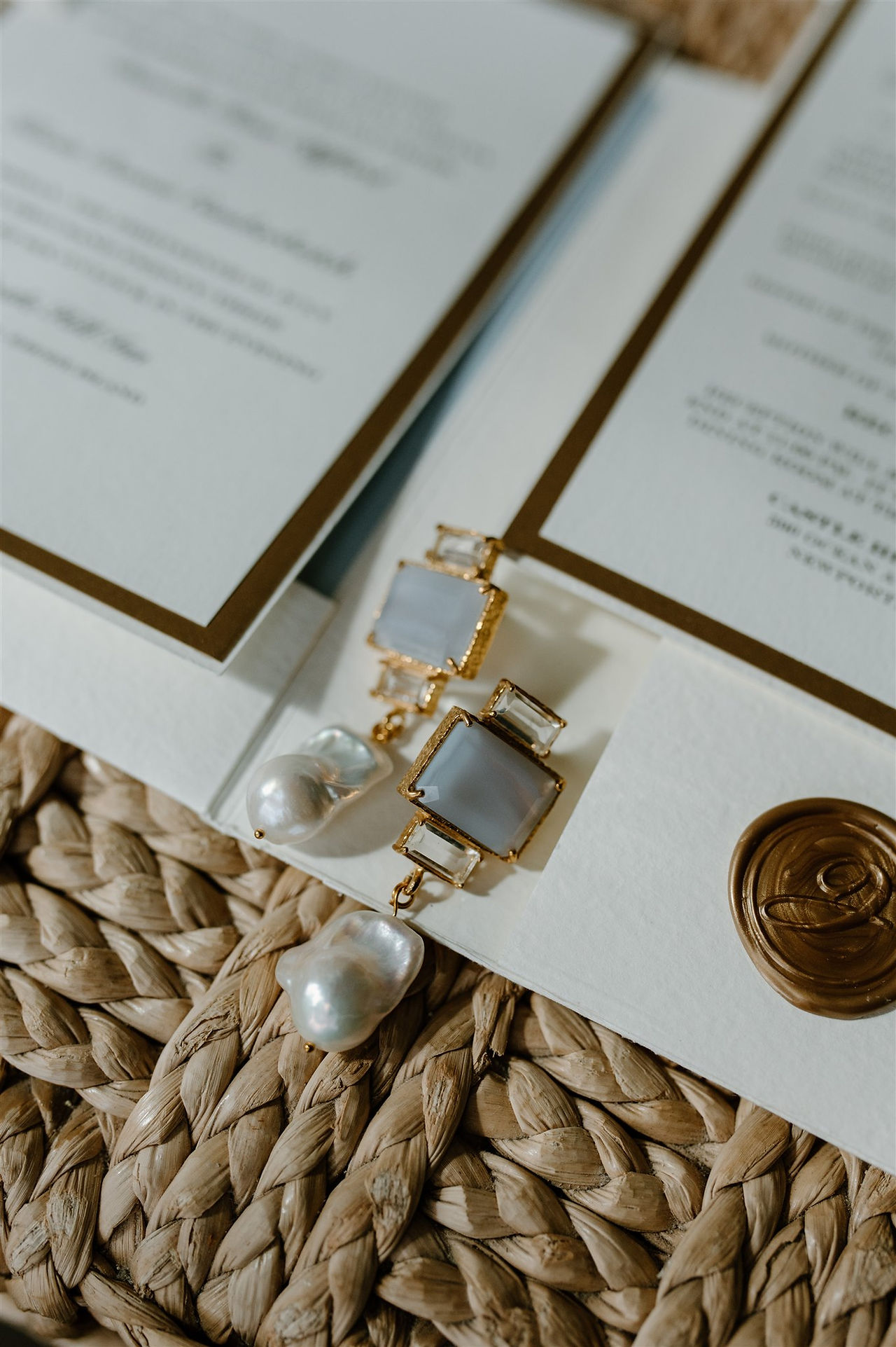 A close-up of an invitation card with gold-framed rectangular and teardrop-shaped pearl earrings placed on it, set on a woven surface.