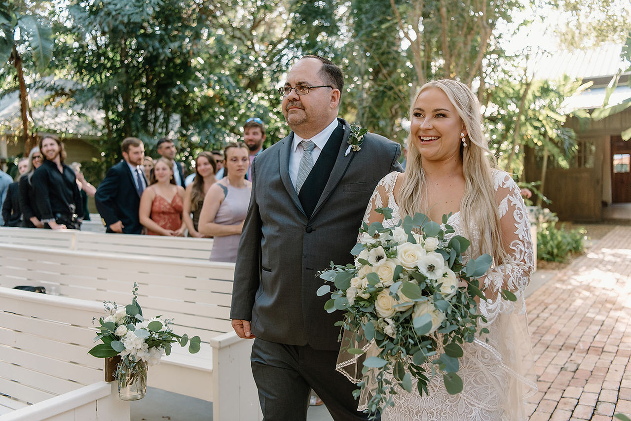 A bride in a white dress holding a bouquet walks with a man in a suit along a garden path in front of a wooden building at the Delamater House