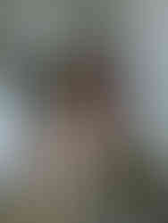 A woman with long hair sits smiling in a chair as a hairdresser styles her hair. Another woman stands in the background. They are in a room with a dress hanging on the door.