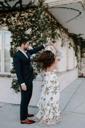A couple walks hand in hand on a sidewalk. The woman is wearing a floral dress, and the man is dressed in a dark suit at their engagement photos in Winter Park