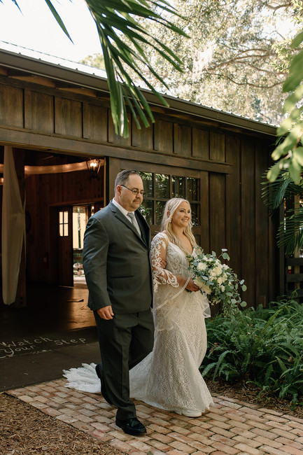 A bride in a white dress holding a bouquet walks with a man in a suit along a garden path in front of a wooden building at the Delamater House