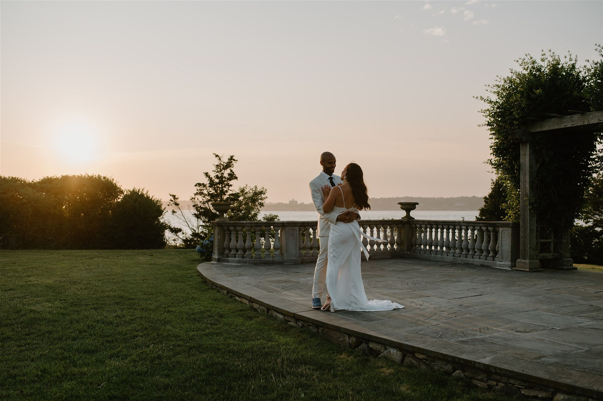A couple dressed in white wedding attire smiles while standing close together, holding a bouquet of white and blue flowers with a serene waterfront backdrop.