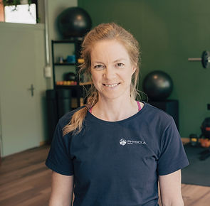 Eine Frau mit rotblonden Haaren steht lächelnd in einem Innenraum. Sie trägt ein dunkles T-Shirt mit Aufdruck "Physiolab". Im Hintergrund sind eine grüne Wand und Fitnessgeräte zu sehen. Die dominierenden Farben sind Grün und Dunkelblau.