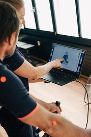 Zwei Personen sitzen vor einem Laptop. Eine Person zeigt auf den Bildschirm, der eine grafische Darstellung anzeigt. Dominante Farben sind Schwarz, Braun und Hellgrau.