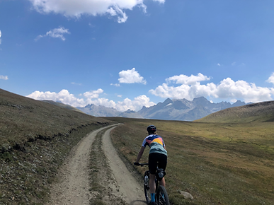 Ein Radfahrer auf einem unbefestigten Weg in einer bergigen Landschaft. Der Himmel ist blau mit weißen Wolken. Dominierende Farben sind Blau und Grün.