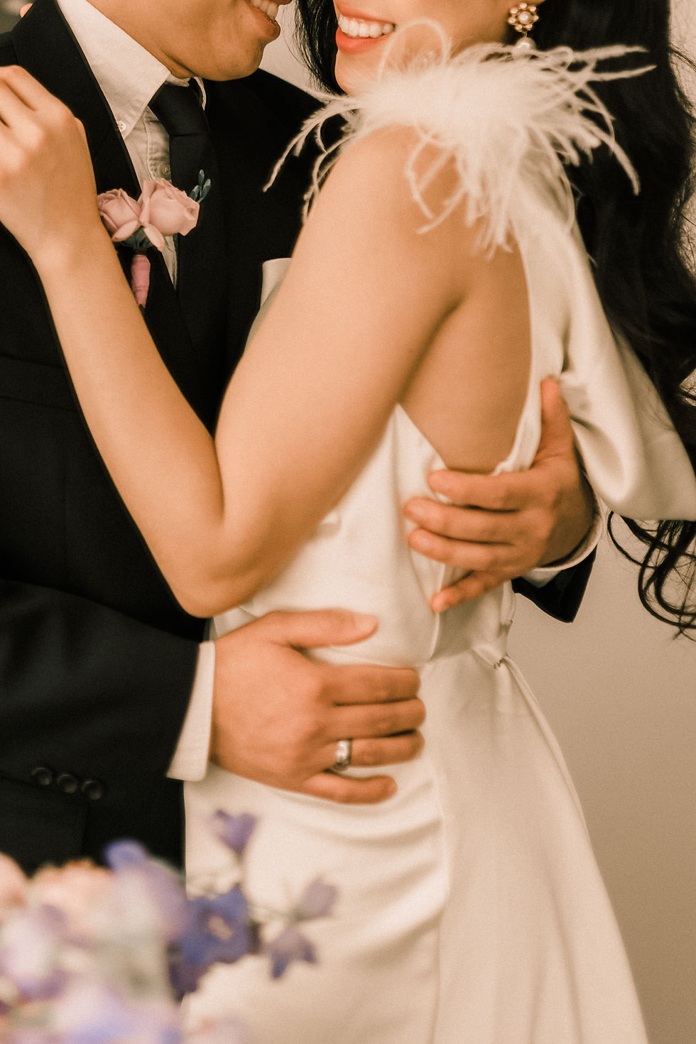 Couple in formal attire embrace, smiling. Man in black suit with boutonnière; woman in white dress with feather detail. Romantic setting.