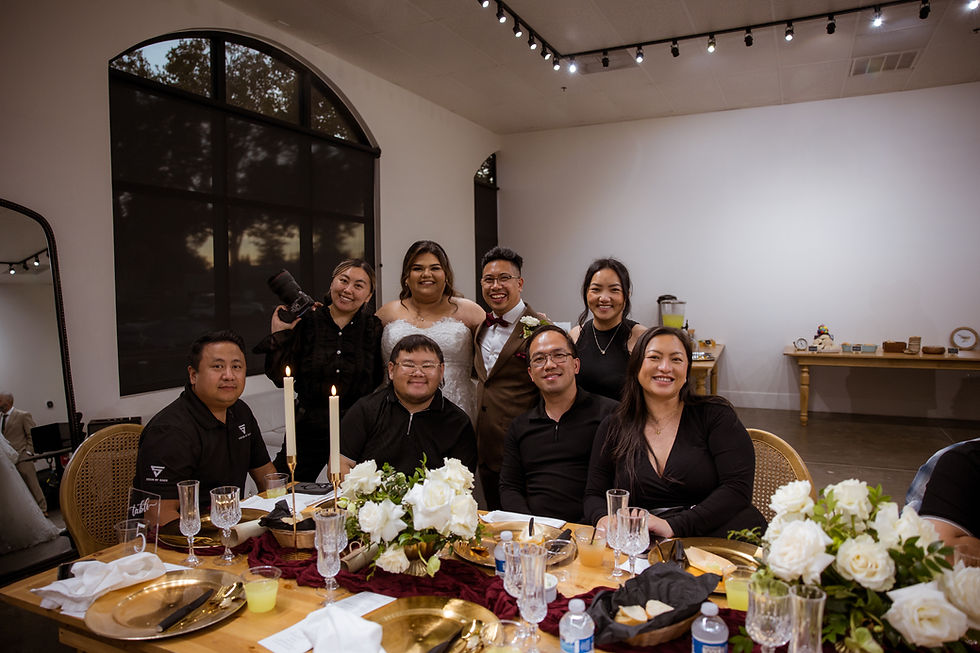 A wedding group photo with smiling people around a decorated table with candles and flowers. A large window and soft lighting set a warm mood.