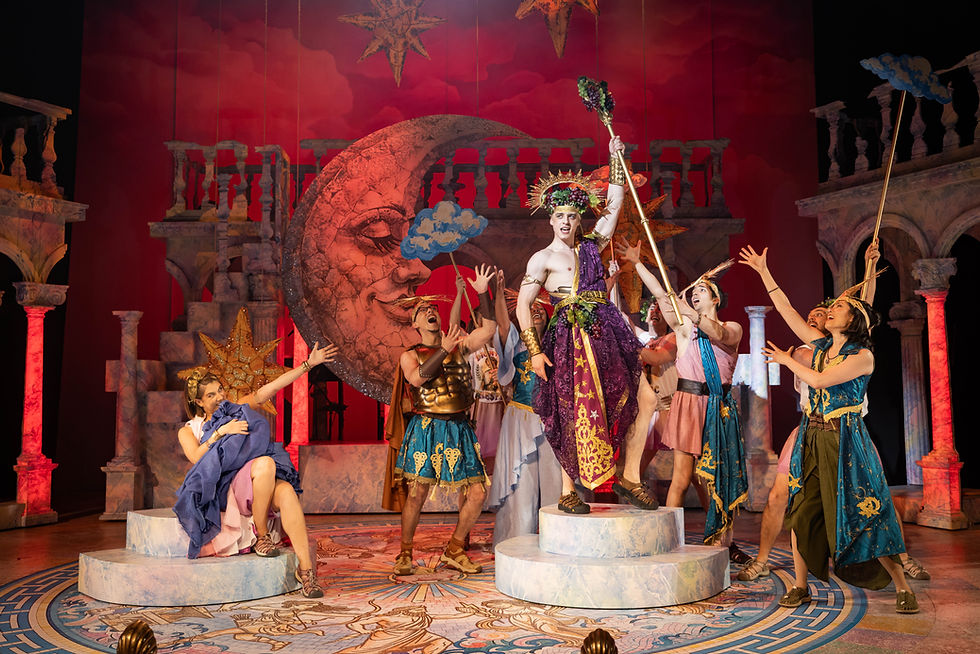 Costumed actors pose on a Greek-style stage, with a central performer in purple holding a staff before a red moon backdrop.