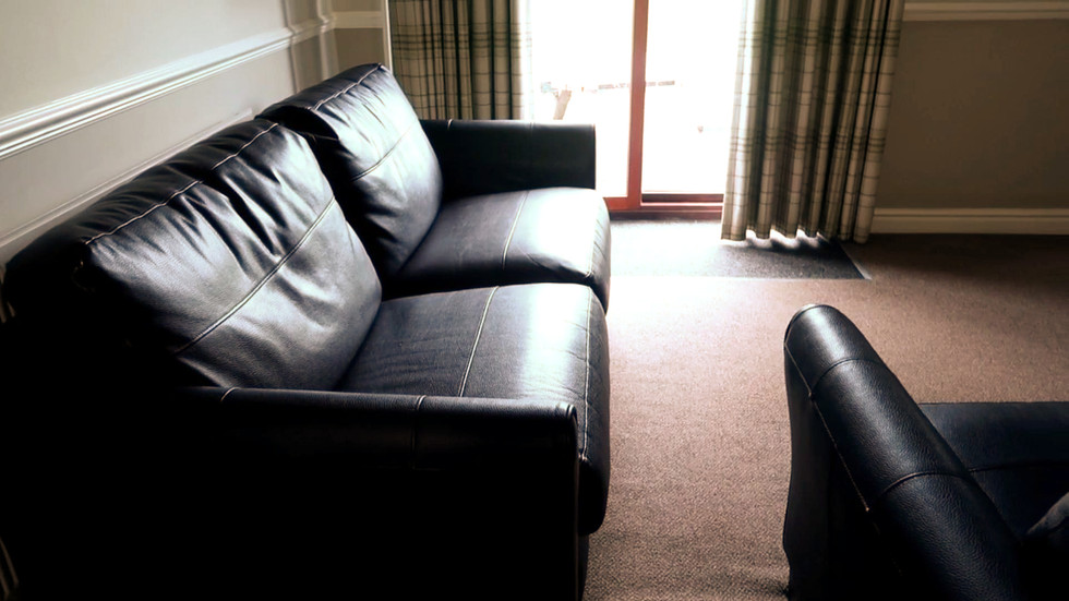 Modern lounge area with black leather sofas, carpeted floor and patio doors leading outside with natural light streaming in.