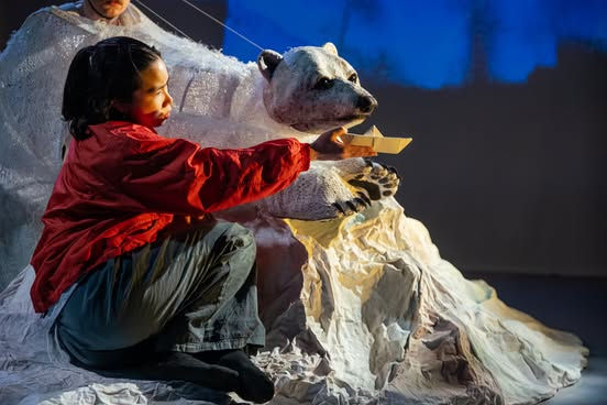 Mika and the Polar Bear - Child in red jacket kneels, feeding a paper boat to a polar bear puppet. Blue-lit background suggests an icy landscape, creating a whimsical mood.