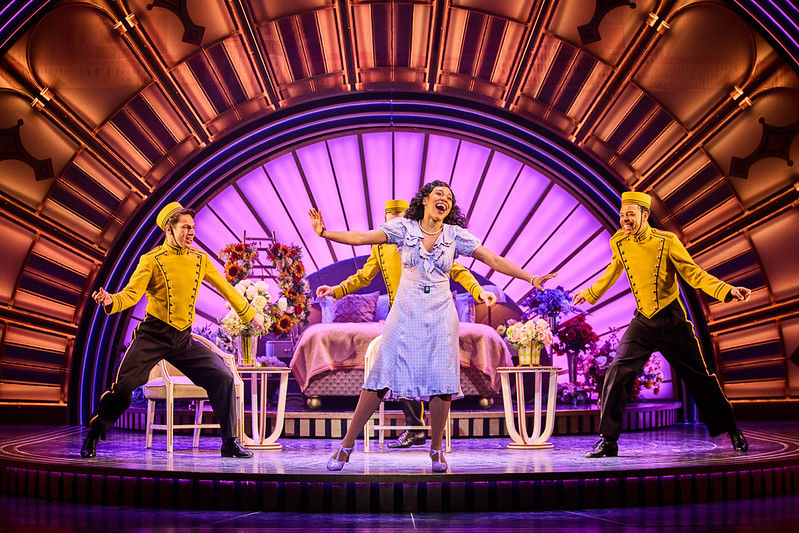 Top Hat the Musical - 
A woman sings joyfully on stage with three men in yellow uniforms. A purple-lit, art deco set with flowers and a bed frames the scene.