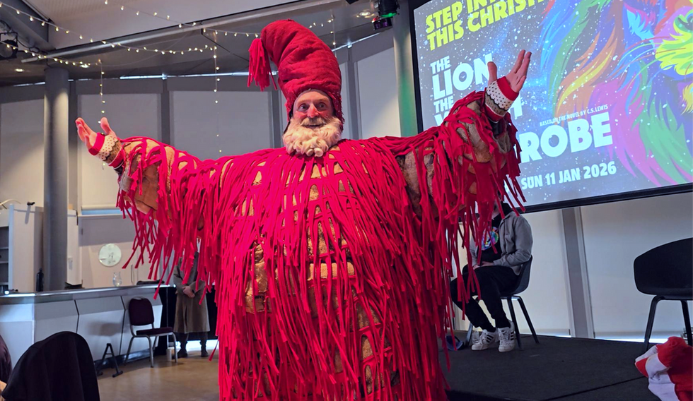 Person in bright red fringed costume poses on stage; screen reads Step Into This Christmas and The Lion, the Witch and the Wardrobe.