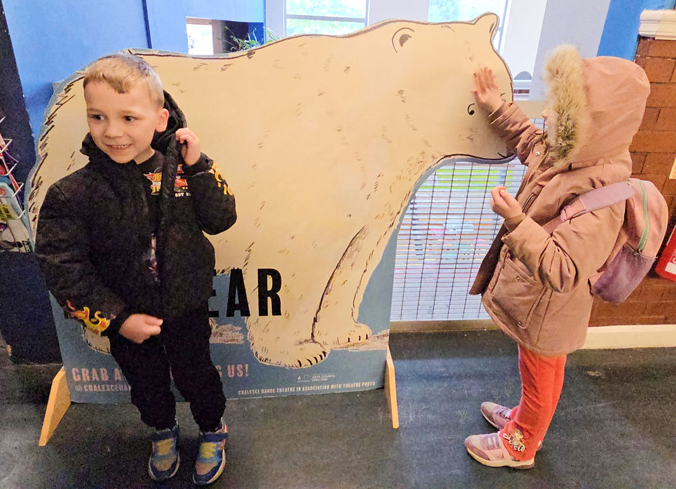 Two children interact with a bear cutout. One boy smiles in a black jacket; a girl in a pink coat and backpack touches the bear's nose.