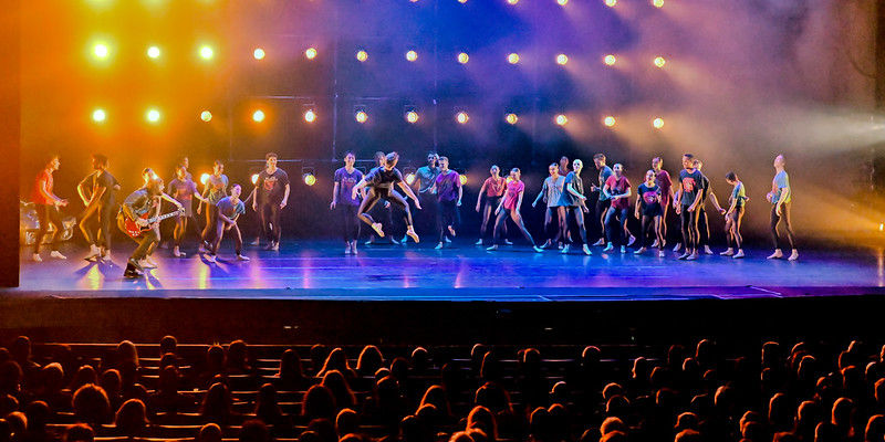 Dancers perform energetically on stage under bright lights with a colorful backdrop. Audience watches in a dimly lit theater.