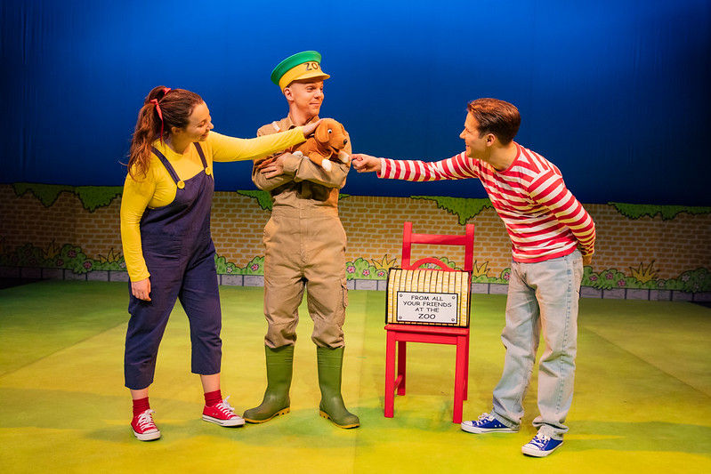 Three actors on a colorful stage share a plush rabbit, beside a red chair with a sign reading FROM ALL YOUR FRIENDS AT THE ZOO.