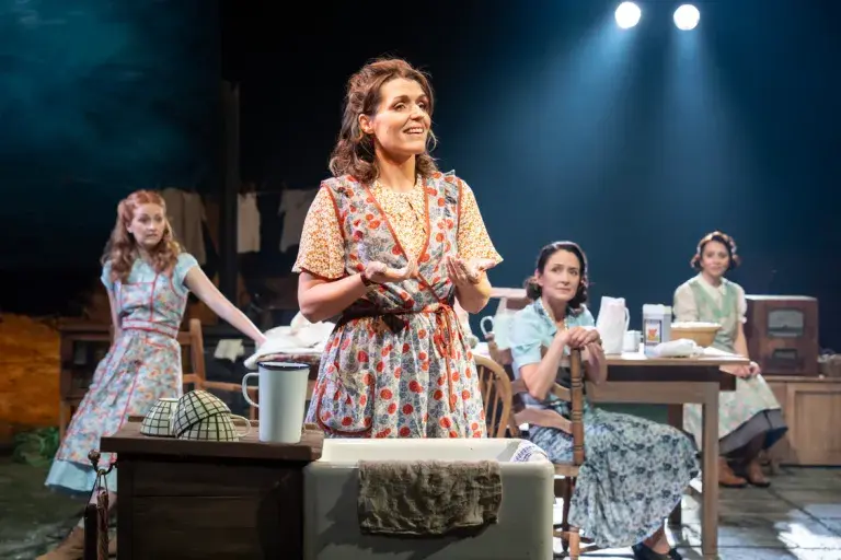Dancing at Lughnasa - Four women in vintage dresses perform on stage. One sings joyfully, surrounded by laundry items, under bright spotlights in a cozy setting.